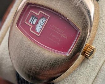 Könnte beinhalten: Eine Lasser Digital Armbanduhr mit einem ovalen Gehäuse in gebürsteter Goldoptik und einem schwarzen, strukturierten Lederarmband. Das Zifferblatt hat einen tiefroten Hintergrund mit einer Digitalanzeige, die die Zahl 10 und eine Minutenscheibe zeigt.