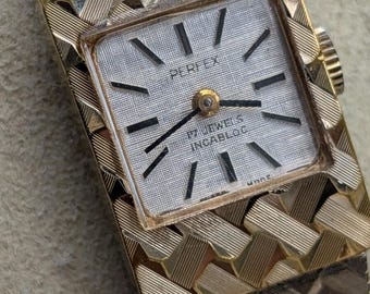 May include: A gold-tone wristwatch with a square face and a textured, woven-link metal band. The watch dial is silver with black baton hour markers and the text Perfex, 17 Jewels, and Incabloc printed in black. A small winding crown is on the side.