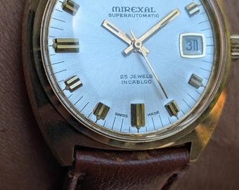 Könnte beinhalten: Eine goldfarbene Mirexal Superautomatic Armbanduhr mit silbernem Zifferblatt, goldfarbenen Stundenmarkierungen und einem Datumsfenster auf der Drei-Uhr-Position. Die Uhr hat ein braunes Lederarmband und trägt die Aufschrift Mirexal Superautomatic 25 Jewels Incabloc.