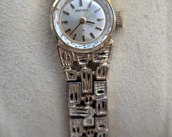 May include: A gold-tone Seiko wristwatch featuring a round silver dial with thin hour markers and a textured, open-link metal bracelet. The watch case has a faceted, reflective bezel, and the word Seiko is printed in black on the dial face.