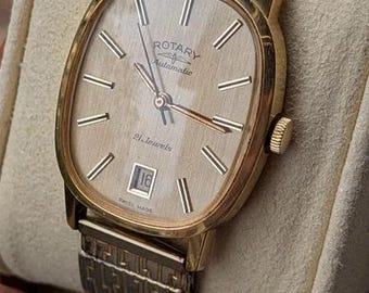 Könnte beinhalten: Eine goldfarbene Rotary Automatikuhr mit strukturiertem champagnerfarbenem Zifferblatt, goldenen Stabindizes und einem Datumsfenster auf der Sechs-Uhr-Position. Die Uhr verfügt über ein Metallgliederarmband mit geometrischem Muster und eine Krone.