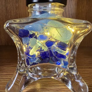 May include: A clear glass bottle with a black lid, filled with blue and green sea glass and tiny fairy lights. The bottle has a unique, flared base and a decorative shape, perfect for home decor.
