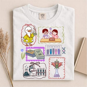 May include: A white t-shirt featuring a colorful graphic print of science-themed illustrations, including a duck in a lab coat, students in a laboratory, books labeled Science Stuff, test tubes, a microscope, and a flower in a flask.