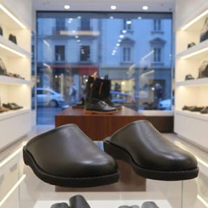 Könnte beinhalten: Ein Paar schwarze Lederclogs mit strukturierter Oberfläche, ausgestellt auf einem Glasregal. Die Clogs haben eine runde Spitze und eine dicke, schwarze Sohle. Weitere Schuhe sind im Hintergrund zu sehen, was auf ein Schuhgeschäft hindeutet.