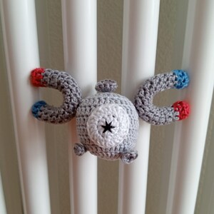 Könnte beinhalten: Handgehäkeltes Magnemite-Plüschtier in Grau, Weiß, Rot und Blau. Das Plüschtier hat eine zentrale Kugel mit sternförmigem Design und zwei gebogene Arme mit farbigen Spitzen. Handgemachtes Spielzeug.