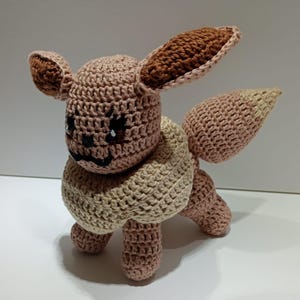 Könnte beinhalten: Ein handgefertigtes Eevee-Plüschtier aus Häkelarbeit. Das Plüschtier hat einen hellbraunen Körper mit dunkelbraunen Ohren und einem cremefarbenen Kragen. Der Schwanz ist in einem helleren Braunton gehalten. Das Spielzeug steht auf vier Beinen.