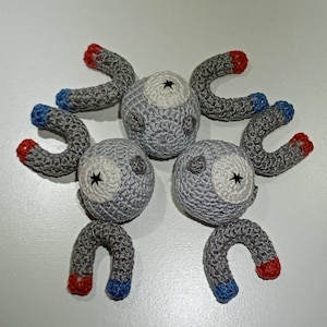 Könnte beinhalten: Gehäkelte Magnemite-Plüschtiere. Jedes Plüschtier hat einen grauen Körper mit einem weißen Auge und einem schwarzen Sterndetail. Die gebogenen Arme sind grau mit roten und blauen Enden. Die Spielzeuge sind auf einer weißen Oberfläche angeordnet.