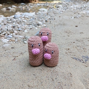 Puede incluir: Tres figuras hechas a mano con ganchillo, que se asemejan a personajes de dibujos animados, se exhiben sobre una superficie arenosa. Cada figura es de color marrón claro con una boca rosa y pequeños ojos negros. Las figuras están de pie, con un arroyo y rocas en el fondo.