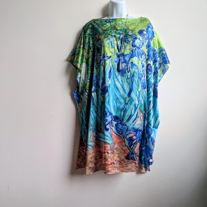 May include: A loose-fitting tunic featuring a vibrant print of blue irises and green foliage on a light background, with a brown earthy tone at the hem. The garment has a wide boat neckline and short, flowing sleeves, paired with a long dark pendant necklace.
