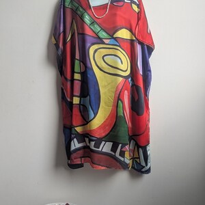May include: A loose-fitting tunic featuring a vibrant, abstract graphic print with bold shapes in red, yellow, blue, green, and purple. The design includes stylized musical instrument motifs and a white pearl necklace draped around the neckline.