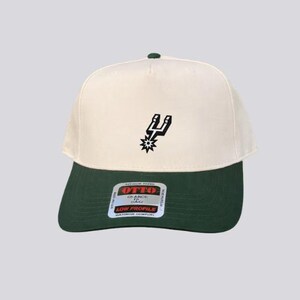 May include: A two-tone baseball cap featuring an off-white crown and a dark green brim. The front panel displays a black and white spur logo. A rectangular sticker on the brim reads OTTO, 65 ANDE, and LOW PROFILE in black and red text.