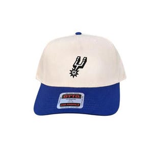 May include: A two-tone baseball cap featuring an off-white crown and a royal blue brim. The front panel displays a black San Antonio Spurs spur logo. A rectangular sticker on the brim reads OTTO CLASSIC FIT MID PROFILE ALL AROUND COMFORT.