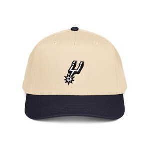 May include: A beige baseball cap with a dark navy blue brim and a small dark navy blue button on top. The front of the cap features a black and white embroidered spur logo centered on the crown. The cap has a structured six-panel design.