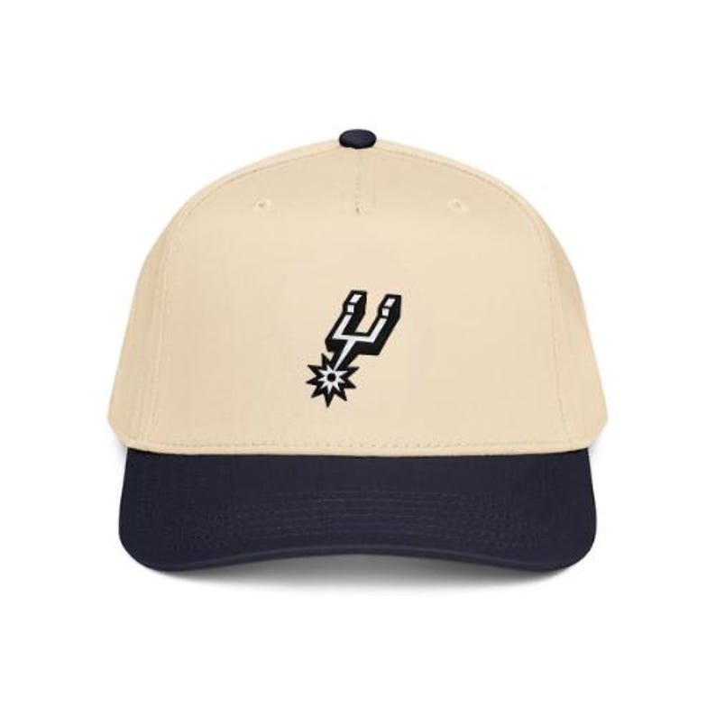 May include: A beige baseball cap with a dark navy blue brim and a small dark navy blue button on top. The front of the cap features a black and white embroidered spur logo centered on the crown. The cap has a structured six-panel design.