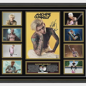 May include: Framed Machine Gun Kelly memorabilia. The display includes a central poster with the artist's image and signature, surrounded by smaller photos. The frame is black with a gold inner border, showcasing a collection of images.