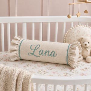 May include: A cylindrical bolster pillow made of beige fabric with ruffled ends and teal piping. The name Lana is embroidered in teal cursive script on the center of the pillow. The pillow is positioned horizontally in a white crib.