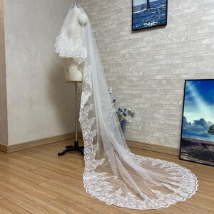 May include: A white bridal veil with detailed lace edging. The veil is draped over a mannequin, highlighting its length and design. The lace trim decorates the edges and top of the veil, creating an elegant look for a wedding.