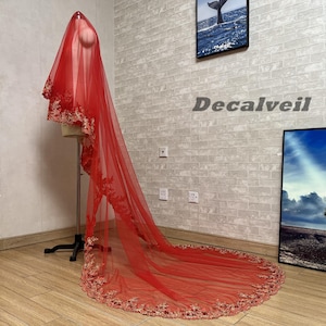 May include: A long, red bridal veil with gold floral embroidery along the edges, displayed on a mannequin. The veil's delicate design is highlighted. The text "Decalveil" is visible on the wall.