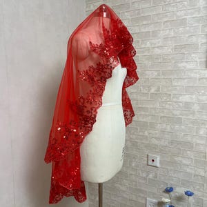 May include: A red bridal veil with a floral lace trim and sequin embellishments. The veil is draped over a mannequin, showcasing its design and texture. The veil is made of a sheer fabric.