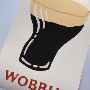 May include: A print featuring a stylized illustration of a pint of stout. The glass is black with a tan head and white foam drips. The word "WOBBLY" is printed in red at the bottom of the image.