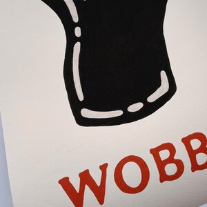 May include: A print featuring a black and white illustration of a pint glass with a creamy head. The word "WOBB" is printed in red, stylized letters at the bottom of the image. The artwork is on a textured, off-white background.
