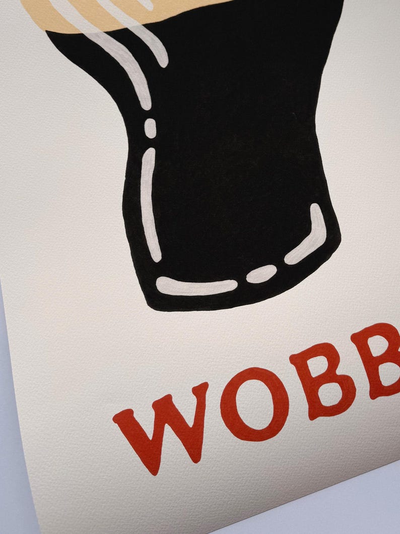 May include: A print featuring a black and white illustration of a pint glass with a creamy head. The word "WOBB" is printed in red, stylized letters at the bottom of the image. The artwork is on a textured, off-white background.