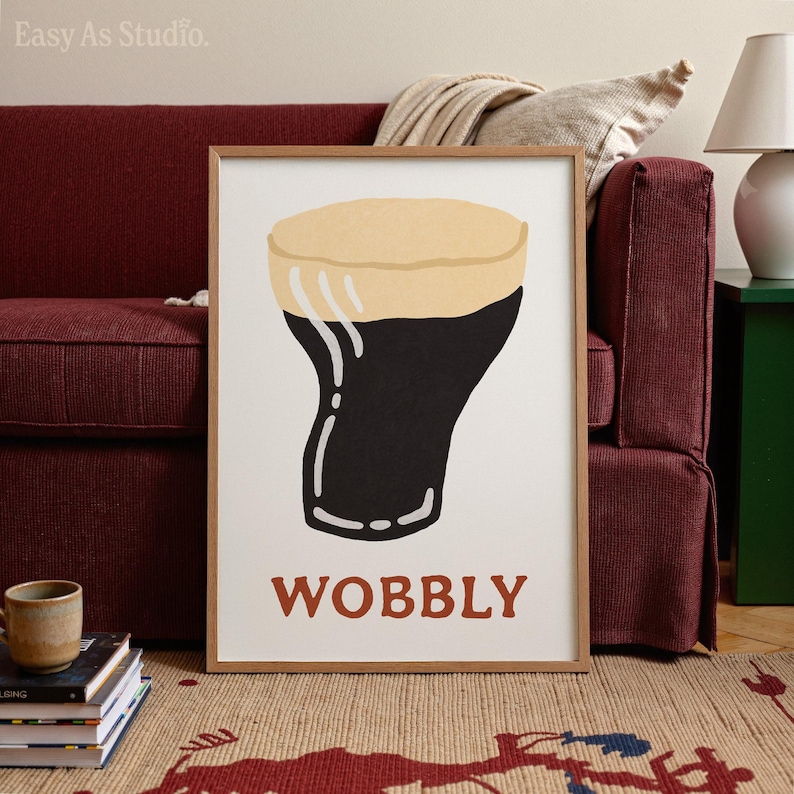 May include: A framed art print featuring a stylized illustration of a pint of stout beer. The beer is dark with a tan head, and the word "WOBBLY" is printed in brown letters below the glass. The print is displayed on a white background.