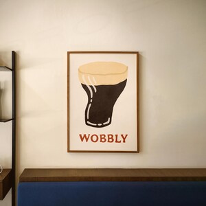 May include: A framed art print featuring a stylized illustration of a pint of stout beer. The artwork has a dark brown body with a tan head, and the word "WOBBLY" in red letters at the bottom.