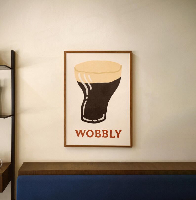 May include: A framed art print featuring a stylized illustration of a pint of stout beer. The artwork has a dark brown body with a tan head, and the word "WOBBLY" in red letters at the bottom.