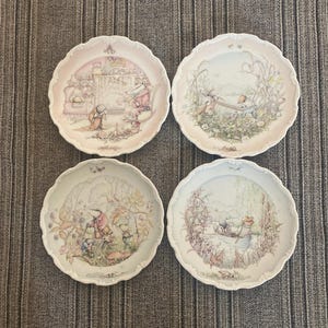 May include: A set of four round ceramic plates with scalloped edges, each featuring detailed illustrations of anthropomorphic animals in various outdoor and indoor settings, rendered in soft pastel colors with intricate line work and decorative borders.