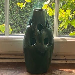 May include: A dark green ceramic vase with a glossy finish and a tapered, organic shape. The vase features multiple circular cutouts distributed across its surface, creating a hollow, sculptural appearance with a narrow opening at the top.