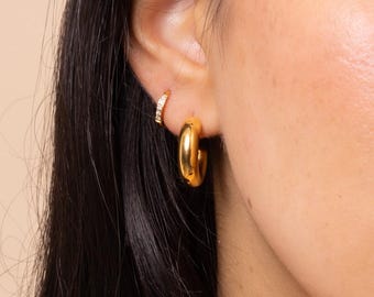 Puede incluir: Primer plano de una oreja adornada con dos pendientes dorados. Uno es un aro pequeño con cristales incrustados, y el otro es un aro dorado más grande, pulido y grueso. Los pendientes están sobre un fondo neutro.
