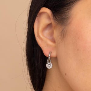 May include: A silver hoop earring with a small, round charm. The charm has a detailed floral design with a central, raised element. The earring is displayed on a person's ear, with the focus on the jewellery.