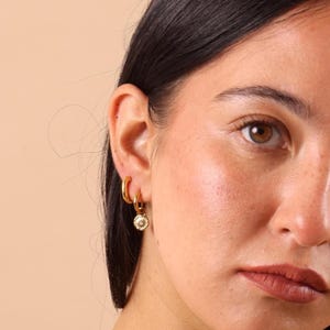 Puede incluir: Pendientes de aro dorados y un pendiente con dije dorado con un pequeño colgante redondo blanco y dorado. Los pendientes se llevan en la oreja de una persona. El fondo es de color beige claro.