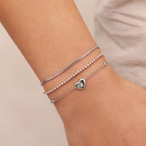 May include: A selection of silver bracelets are displayed on a wrist. The bracelets include a fine chain, a row of small, sparkling stones, and a bracelet with a heart-shaped charm. The heart charm features a small, sparkling stone at its centre.
