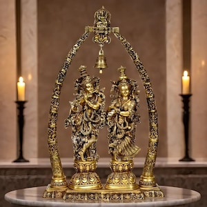 May include: A golden statue depicting two figures, one playing a flute, under an ornate arch. The figures stand on detailed pedestals, with a small bell hanging from the arch. The statue is set against a neutral background with lit candles on either side.