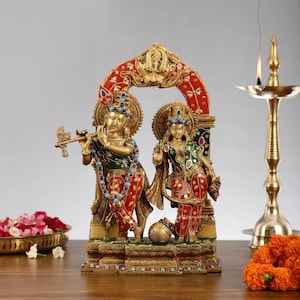 Può includere: Una statua dorata raffigurante due figure, una che suona un flauto. Le figure sono adornate con abiti colorati con accenti rossi, verdi e blu. La statua è ambientata contro un arco decorativo con dettagli rossi e dorati.