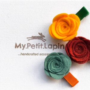 May include: Three felt flower hair clips in yellow, burgundy, and teal. Each flower is attached to a ribbon-covered barrette in green, white, and orange. The logo "My.Petit.Lapin" is printed on the white background.