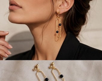 May include: A pair of gold-toned drop earrings featuring an elongated oval frame. Each earring is suspended from a small gold ball stud and contains two small, smooth black beads positioned along the vertical wire of the oval-shaped metal frame.