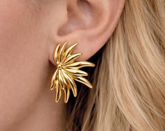 May include: A gold-colored earring with a floral design is shown. The earring is a cluster of curved, pointed shapes radiating from a central point. The earring is worn on an ear, with blonde hair visible.