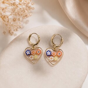 May include: A pair of gold-toned hoop earrings featuring heart-shaped pendants. Each pendant contains a clear center filled with small, colorful floral-shaped accents in shades of orange, yellow, white, and blue, suspended from a circular hoop.