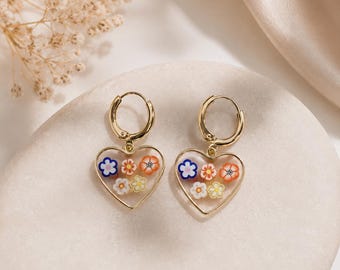 May include: A pair of gold-toned hoop earrings featuring heart-shaped pendants. Each pendant contains a clear center filled with small, colorful floral-shaped accents in shades of orange, yellow, white, and blue, suspended from a circular hoop.