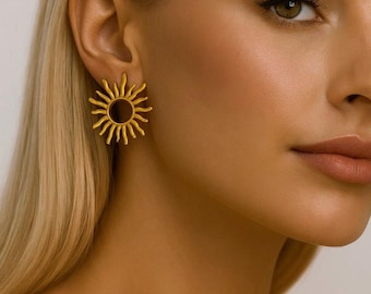 May include: Gold-colored sunburst earrings. Each earring features a circular center with radiating rays, creating a sun-like design. The earrings are worn on a person's ear, adding a touch of celestial charm to the overall look. These earrings are a stylish accessory.