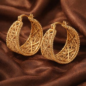 May include: A pair of gold-colored hoop earrings. The earrings have an intricate filigree design with a beaded border. The earrings are displayed on a brown satin fabric.