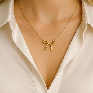 May include: A gold-colored necklace featuring a bow pendant. The bow is centered on a delicate chain, creating a minimalist and elegant design. The necklace is worn against a white collared shirt.