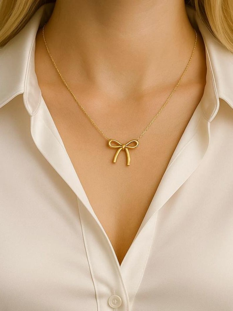 May include: A gold-colored necklace featuring a bow pendant. The bow is centered on a delicate chain, creating a minimalist and elegant design. The necklace is worn against a white collared shirt.