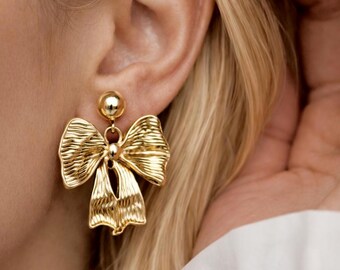 May include: Gold-colored bow earrings. The earrings feature a textured bow design with a small ball at the top. The earrings are hanging from a person's ear, with blonde hair and a white shirt in the background.