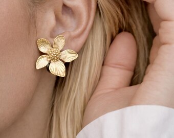 May include: Gold-colored flower-shaped stud earrings. The earrings have detailed petals and a textured center, resembling a blooming flower. The earrings are worn on an ear, with blonde hair and a white shirt in the background.