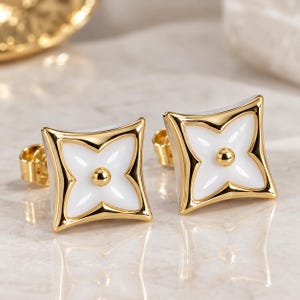 May include: A pair of gold stud earrings featuring a square shape with concave sides and a white enamel inlay. Each earring displays a central gold sphere surrounded by a four-petaled floral motif, finished with a polished gold metal border.