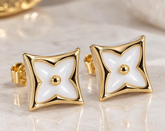 May include: A pair of gold stud earrings featuring a square shape with concave sides and a white enamel inlay. Each earring displays a central gold sphere surrounded by a four-petaled floral motif, finished with a polished gold metal border.